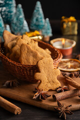 Close-up of basket with gingerbread cookies on table with cinnamon, star anise, orange, trees and candle, black background, vertical, with copy space