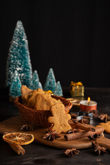 Top view of basket with gingerbread cookies on table with cinnamon, star anise, orange and Christmas trees, black background, vertical, with copy space