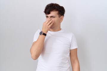 Young non binary man wearing casual white t shirt smelling something stinky and disgusting,...