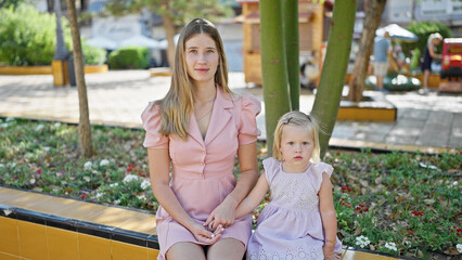 Fototapeta premium Caucasian mother and daughter, serious faces etched in concentration, sitting together on a park bench, sharing an emotive moment amidst the green outdoors.