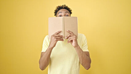 African american man covering mouth with book over isolated yellow background