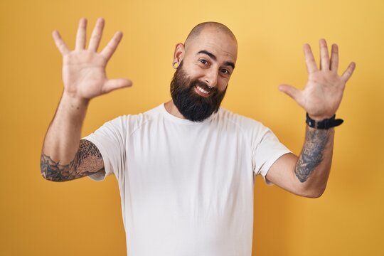 Young hispanic man with beard and tattoos standing over yellow background showing and pointing up with fingers number ten while smiling confident and happy.
