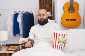 Young hispanic man with beard and tattoos eating popcorn in the bed clueless and confused...