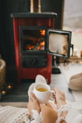 Young woman sitting by the fireplace in white sweater, drinking tea in cozy log cabin.