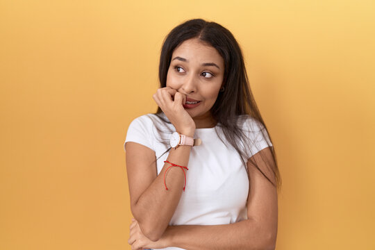 Young arab woman wearing casual white t shirt over yellow background looking stressed and nervous with hands on mouth biting nails. anxiety problem.