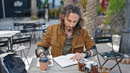 Young hispanic man tourist reading book sitting on table taking notes at coffee shop terrace