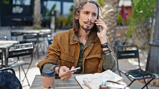Young hispanic man tourist talking on smartphone holding credit card sitting on table at coffee shop terrace
