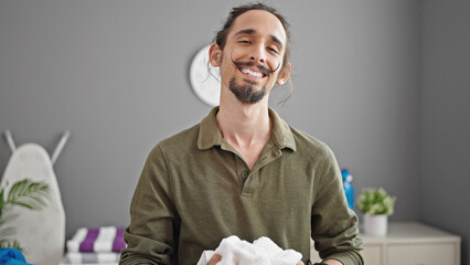 Young hispanic man smiling confident holding towel at laundry room