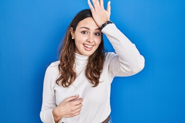 Young hispanic woman standing over blue background surprised with hand on head for mistake, remember error. forgot, bad memory concept.