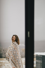 Fototapeta premium Young woman wrapping in a warm blanket standing on a balcony with snowy forest on the background.