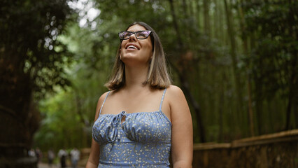 Naklejka premium Beautiful hispanic woman with glasses, smiling confidently as she stands amongst kyoto's lush bamboo forest, looking around, full of joy and amazement.
