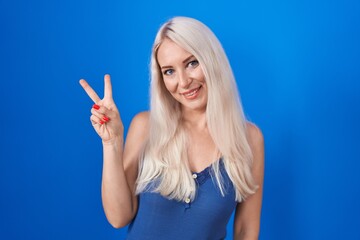 Caucasian woman standing over blue background smiling with happy face winking at the camera doing...