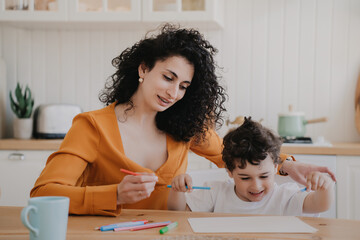 Creative little curly boy drawing at home sitting with mom at desk at kitchen. Family domestic leisure, childhood. Cheerful young hispanic woman spending time with son. Babysitter with kid.