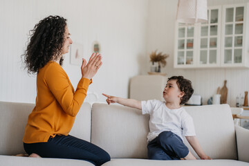 Your turn! Curly young Italian woman teaching son to give five. Caucasian babysitter in orange blouse playing with kid at kitchen sitting on sofa clapping palms. Elder sister entertaining cute brother - Powered by Adobe