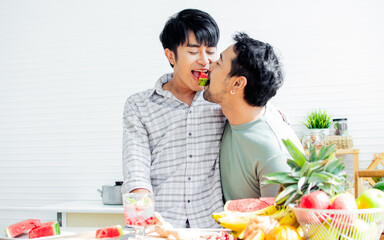 Gay LGBT sweet Asian couple wearing pajamas, smiling, bite, eating piece of watermelon together with happiness,  love while having breakfast in kitchen at home in the morning. Lifestyle Concept.