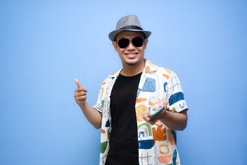 asian man wearing beach shirt holding a phone while pointing a finger standing over blue background. Vacation concept