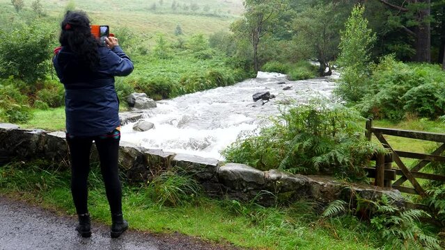 Woman standing on the stone bridge over streaming mountain river and taking pictures using her smartphone. When finished turns around and smiling walks away from beautiful scene in Lake District.