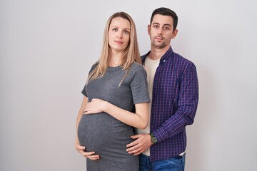 Young couple expecting a baby standing over white background relaxed with serious expression on...
