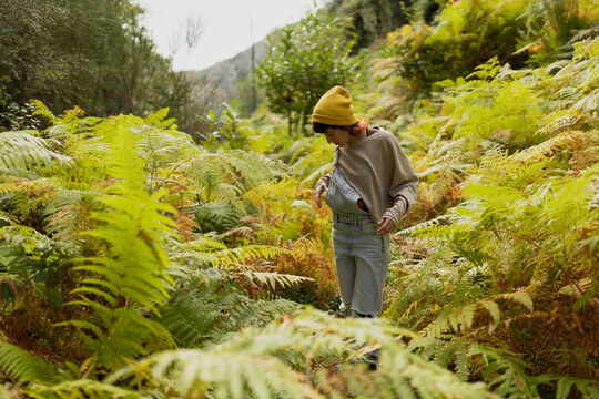 Woman with mobile phone and headphones in fern forest wearing yellow hat, listening to music, taking photos and video call