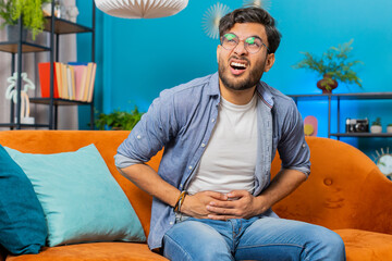 Indian Arabian man sits on sofa feeling sudden strong abdominal stomach ache gastritis problem. Bearded Arabian guy having symptom poisoning diarrhea indigestion peptic ulcer pancreatitis at home room