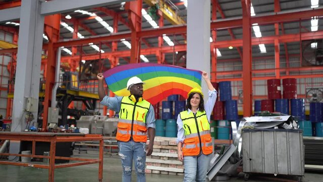 Workers Celebrate Pride Month in Industrial Setting: Two diverse workers, a man and a woman, stand proudly in a large industrial warehouse.
