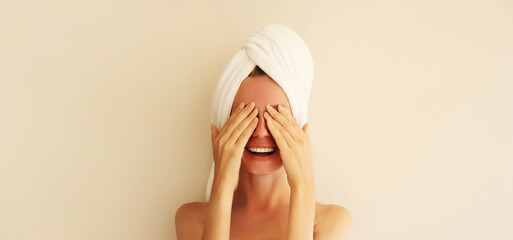Natural beauty portrait of happy smiling young caucasian woman touches her clean skin, covers her eyes while drying wet hair with white wrapped bath towel on her head after shower