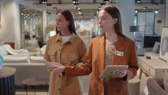 Furniture store manager walking alongside with client while showing her various mock-ups and models available in store