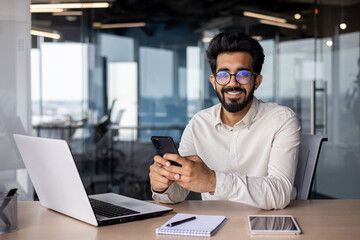 Portrait of a young Indian businessman, office worker sitting at a desk and talking on the phone, smiling and looking at the camera