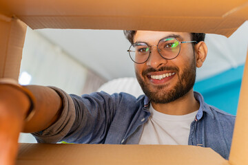 View from inside the box. Happy Indian man unpacking delivery parcel at home. Smiling satisfied guy...