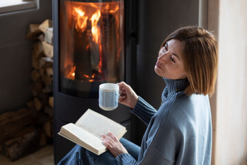 Young attractive woman sitting close to fireplace, reading interesting book and drinking hot tea in the cozy house