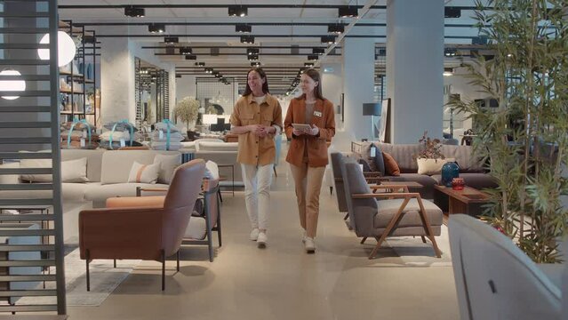 Full shot of saleswoman with badge and female customer walking around furniture store, looking out necessary pieces