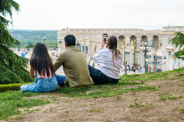 Three friends sit on grass overlooking city square, with one capturing the moment on a phone