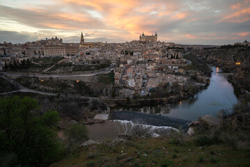 Historic city of Toledo with the a weir of Tagus River in foreground from a viewpoint of the city at sunset with reddish sky. UNESCO World Heritage Site - obrazy, fototapety, plakaty
