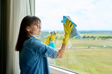 Middle-aged woman in gloves with spray cleaning windows at home