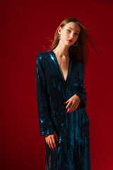 Fototapeta premium Fashionable confident woman wearing trendy blue sequin v-neck party dress, posing on red background. Studio fashion portrait. Copy, empty space for text