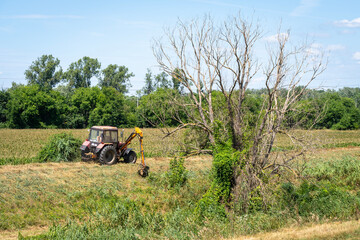 Lawn tractor working on a stream bank next to a dead tree in the summer sun