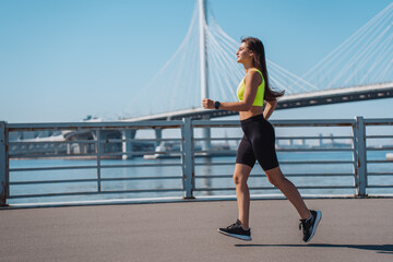 Young hispanic brunette woman in sportswear running at embankment  against huge bridge on background. Fit Caucasian girl practicing sport outdoors at sunny summer day. Successful female on workout.