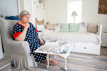 An elderly woman in thoughts while collecting puzzle while sitting in a chair in her bright room at home. - obrazy, fototapety, plakaty