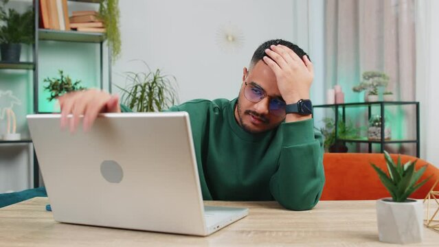 Displeased sad Indian man use laptop notebook typing browsing working, loses becoming surprised sudden lottery results bad news fortune loss game fail computer virus. Arabian guy at home office table