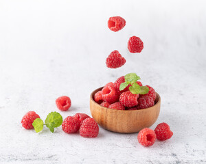 Fresh sweet raspberry falling into wooden bowl with berries and mint leaves on light stone...