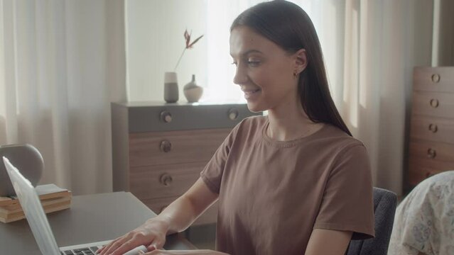 Medium shot of Caucasian young woman sitting in her studio behind desktop, remotely working on laptop