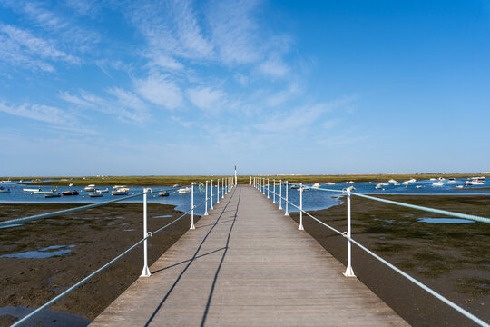 Empty walkway over blue ocean, coastline and horizon seen a large long wooden jetty with a thin white metal railing along the edges but in the perspective distance a blue sky and an ocean with boats.