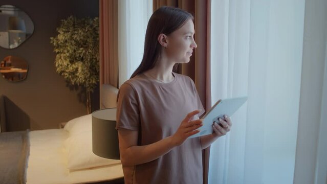 Woman in comfortable home clothes standing in front of window of her living room, scrolling through social media on digital tablet