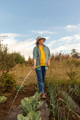 Mujer latina adulta caminando con una manguera llegando las hortalizas en medio de su huerta foto...