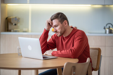 Sad frustrated man procrastinating after fired from job looking at laptop screen on kitchen at...