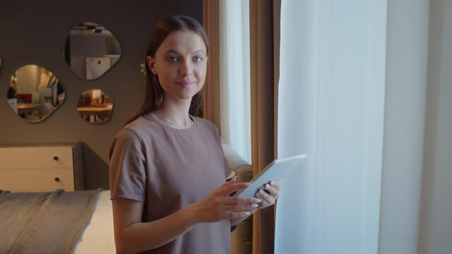 Portrait of young woman in beige t-shirt standing next to window of her studio, looking at digital tablet, then at camera
