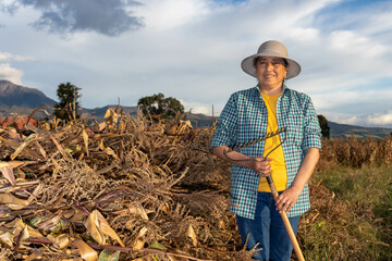Mujer latina parada en frente de cañas de maiz cortadas y de fondo un silo, granero y las montañas esta sonriendo sosteniendo su rastrillo 