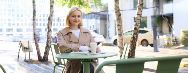 Image of young woman, makes sketches outdoors on street in notebook, drinks coffee and smiles at...