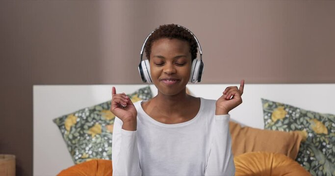 Portrait of delighted cheerful african american dark skinned woman wearing headphones enjoying favourite songs listening to music dancing gesturing relaxing at weekends after hard work days .
