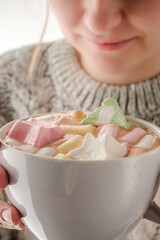 A woman is holding a mug with hot cocoa decorated with marshmallows on the background of a winter sweater. Chocolate bomb.